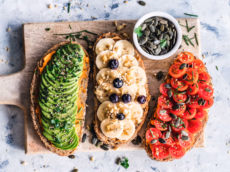 Three slices of toast with avocado, banana, blueberries, and tomatoes