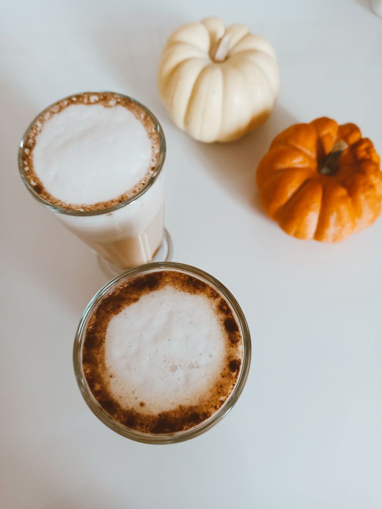 Top-down view of two frothy pumpkin spice lattes and autumn pumpkins on a white surface.